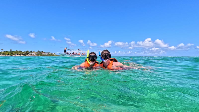 Two snorkelers at the surface of Akumal Bay, with the palm-lined shore and a tour boat behind them